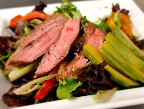 Close up of sliced cooked flank steak resting on a bed of vegetables and salad greens.