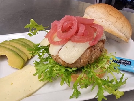 Open-faced turkey burger on a bun topped with pink pickled onions and two slices of white cheese. The sandwich is on a cutting board paired with avocado slices, white cheese and frilly, green lettuce leaves.