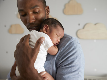 A man snuggles a newborn infant against his chest standing in front of a wall with clouds on it.