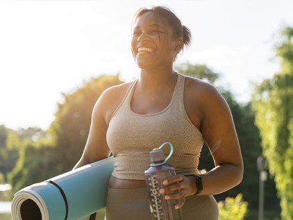 Smiling woman carrying a yoga mat and water bottle on a sunny day