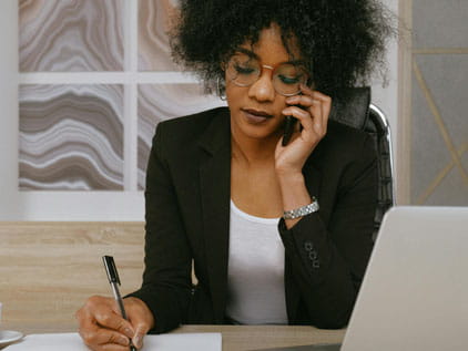 Medium close up of a young woman in a dark colored sweater sitting in a beige-colored office with a phone to her ear while sitting at a desk and taking notes.